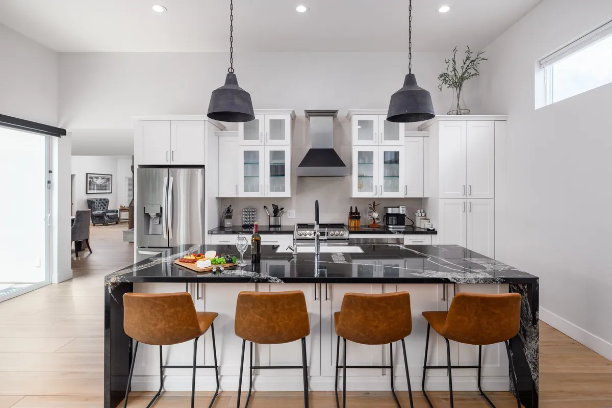 A contemporary kitchen scene featuring an island with a striking black marble countertop, surrounded by stylish bar stools. Pendant lights hang above the island, casting a warm glow over the inviting space. Stainless steel appliances line the back wall, and the arrangement encourages both casual dining and lively conversation while cooking or entertaining.