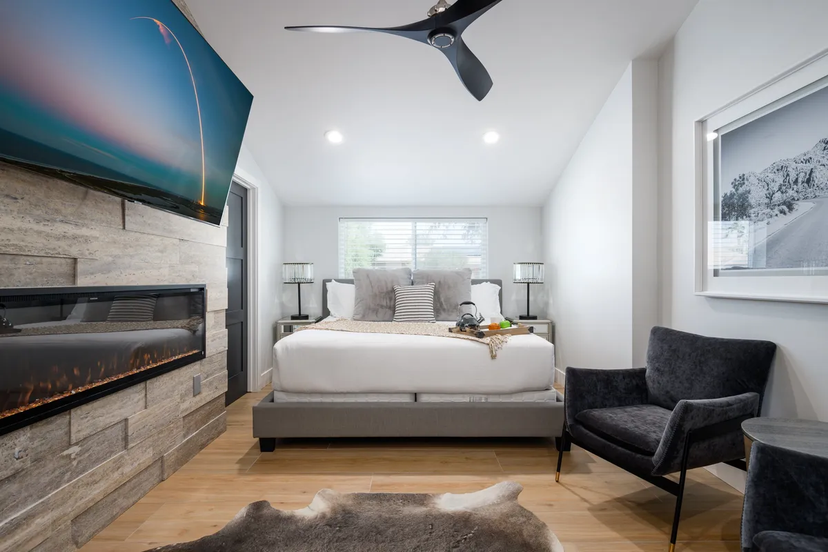 A beautifully appointed primary suite featuring a plush bed with decorative pillows, a modern fireplace, a large flat-screen TV, and a cozy armchair. Elegant wall art and soft lighting enhance the tranquil atmosphere, while the space feels inviting for relaxation and privacy.