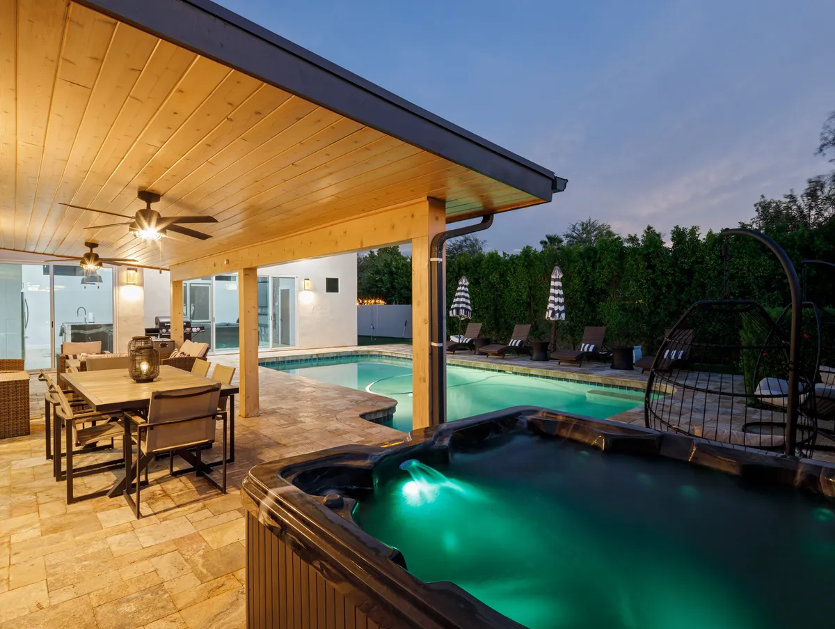 An outdoor dining area set beside a tranquil swimming pool, featuring a table with comfortable seating for group meals and lounge chairs nearby. The backdrop includes a hot tub and lush landscaping, creating a serene setting for al fresco dining under the stars or intimate gatherings by the water.