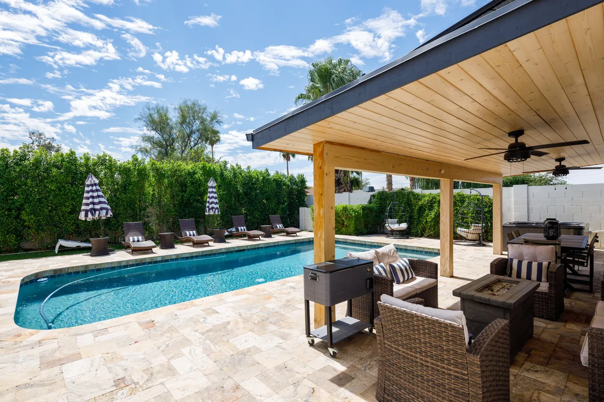 A serene exterior space featuring a swimming pool, lounge chairs, a covered patio, and stylish outdoor furnishings. The area is designed for sunbathing, dining, and evening gatherings, evoking a tranquil, resort-inspired mood. A fire pit is visible, hinting at cozy evenings under the stars.