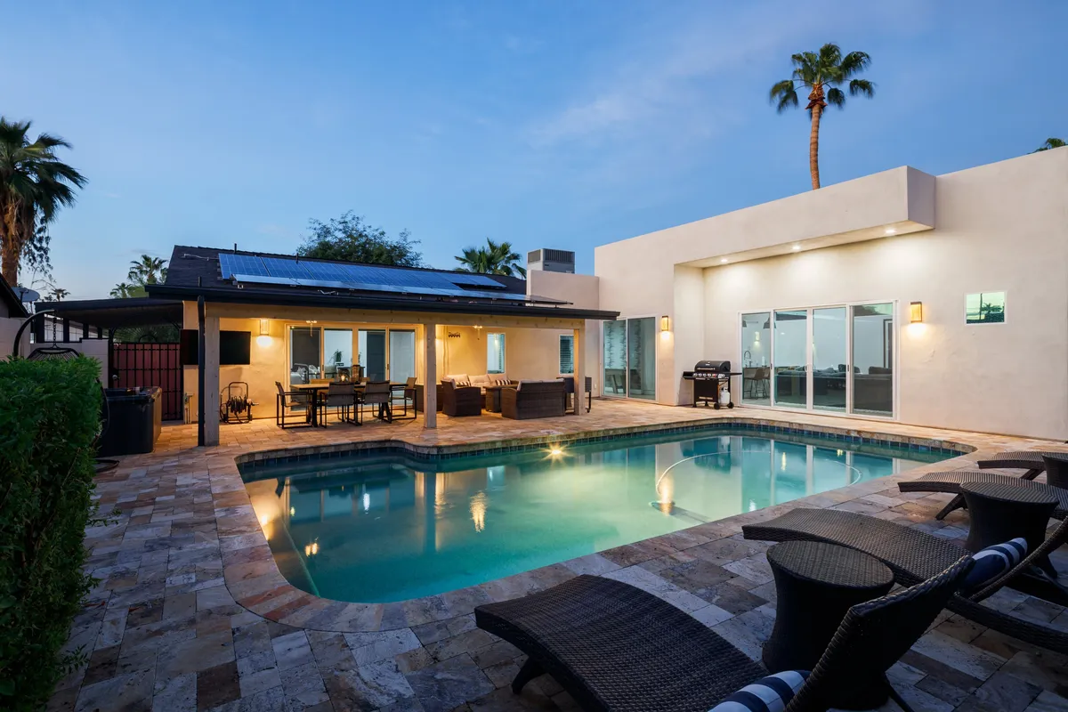 The image showcases the property's inviting backyard oasis at sunset, featuring a sparkling swimming pool surrounded by lush palm trees, a modern outdoor seating area, and a spacious patio equipped with solar panels. The scene is set for relaxing afternoons, lively poolside gatherings, and intimate al fresco dining under the Arizona sky.