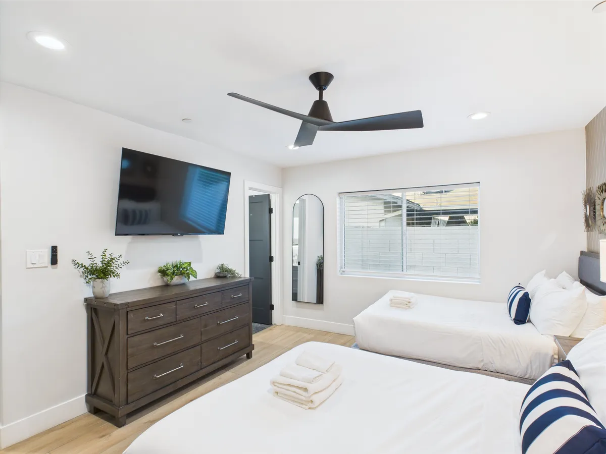 A modern secondary bedroom with two beds, a large window bathing the room in natural light, a TV, a dresser, and a decorative mirror. The layout suggests both comfort and flexibility for guests, accommodating multiple people with ease and style.