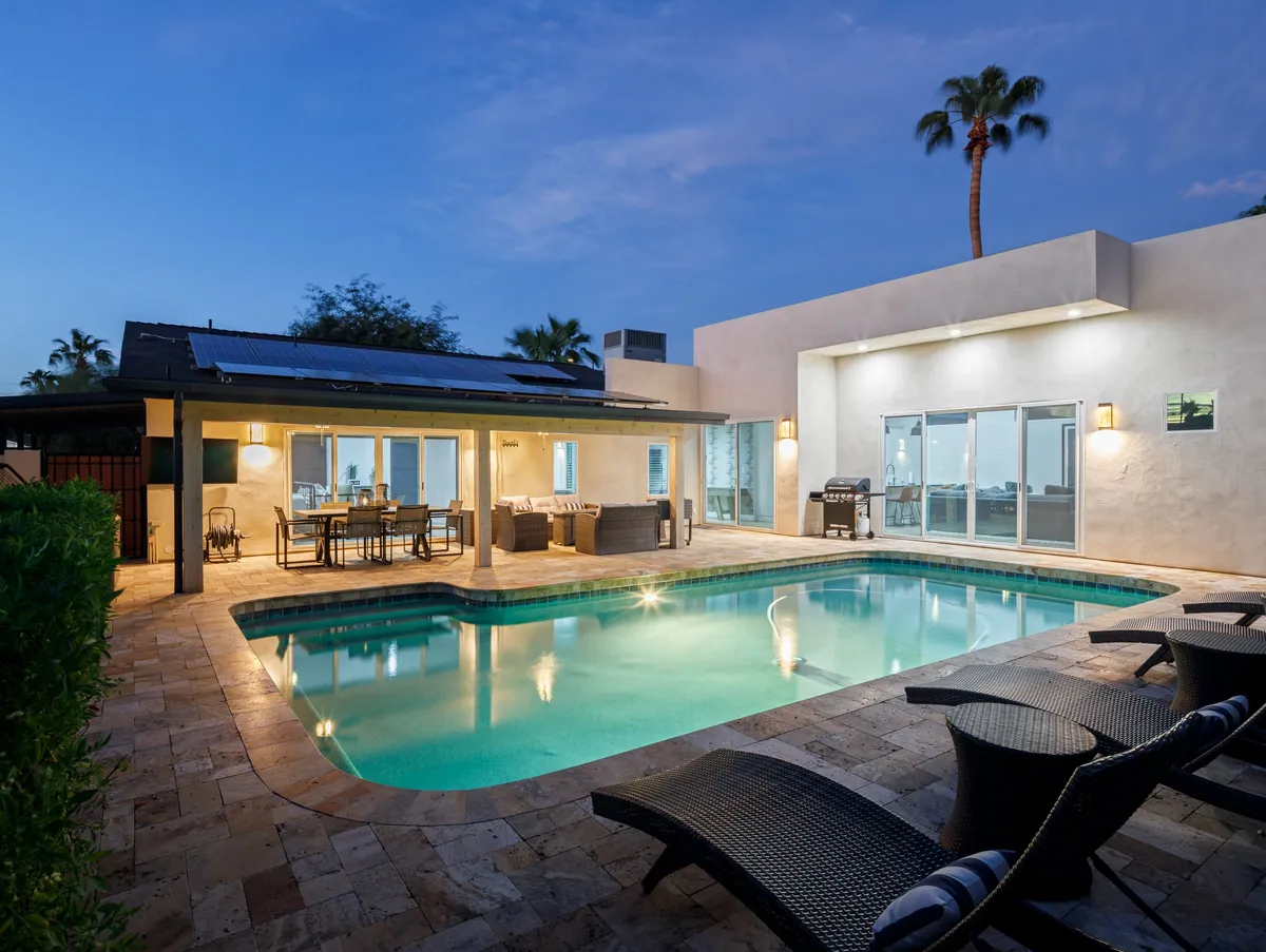 A tranquil, luxurious backyard scene featuring a sparkling swimming pool surrounded by palm trees and an expansive outdoor seating area. The view through sliding glass doors hints at seamless indoor-outdoor living, with spaces for swimming, sunbathing, barbecuing, and relaxing together.