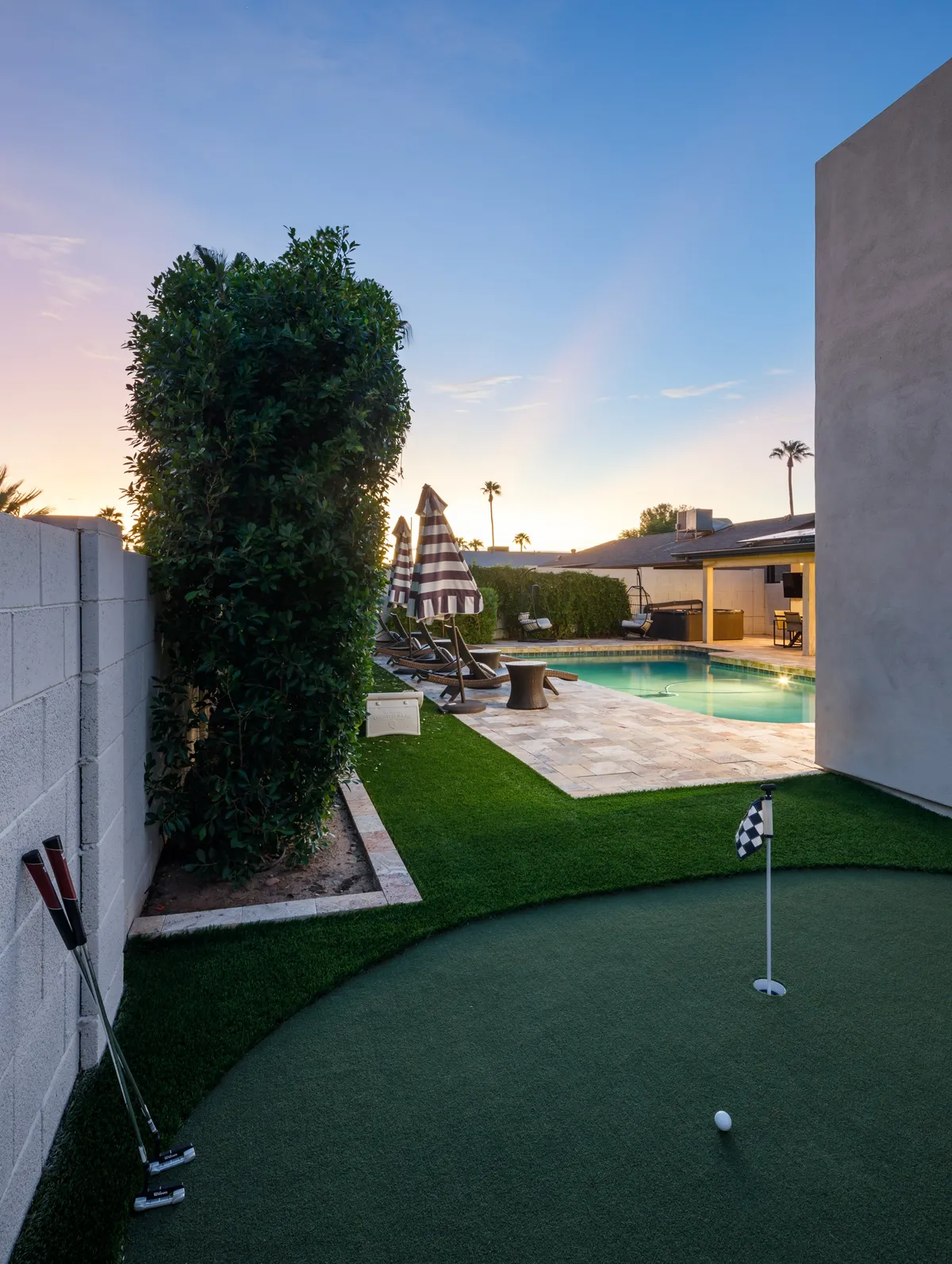 A sunny exterior scene featuring a private putting green, a sparkling swimming pool, several lounge chairs under an umbrella, and lush landscaping. The setting feels leisurely and is clearly designed for both golf practice and poolside relaxation.