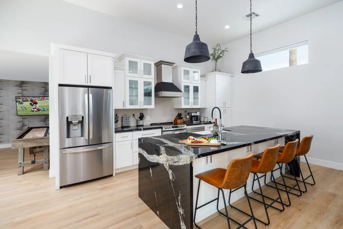 A bright, modern kitchen with a large island at its center, surrounded by sleek stainless steel appliances. Four bar stools line the island, inviting guests to sit and chat while meals are prepared. A wall-mounted TV is visible, adding a touch of entertainment to the culinary hub. The open layout and ample counter space convey an atmosphere designed for cooking, entertaining, and gathering.